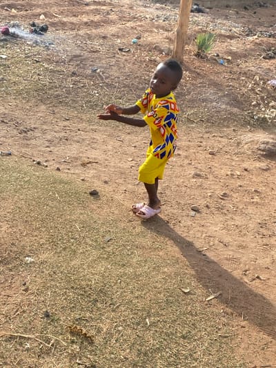 A young child stands on an uneven, light brown dirt ground interspersed with dry grass in an outdoor area of Rido, Nigeria. The child, identifiable as having dark skin and short dark hair, is clad in a yellow short-sleeved shirt featuring blue, red, and white diamond patterns, yellow shorts, and light pink open-toed slides. The child's body is slightly angled, with the head tilted upwards and to the left, and both hands are raised approximately to chest height, palms open and fingers slightly curled, indicating an ongoing action or gesture. A distinct, elongated shadow extends from the child's feet towards the lower right of the frame, suggesting a low sun angle. In the upper left background, a small accumulation of dark debris emits a faint wisp of smoke. A slender, light-colored wooden post or tree trunk is visible vertically behind the child to the right. The ground contains scattered small stones, dry organic matter, and various small pieces of litter.