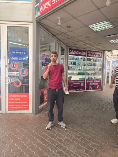 A man is standing in front of a store in Tbilisi, Georgia. He is wearing a red t-shirt and dark pants. The man is holding a cigarette to his mouth and appears to be smoking it. The store is a Samsung store, and it sells electronics. The store is located on a street with cobblestone pavement. The man is the only person clearly visible in the foreground. The store has large glass windows displaying various electronic devices. The overall environment appears to be an urban commercial area.