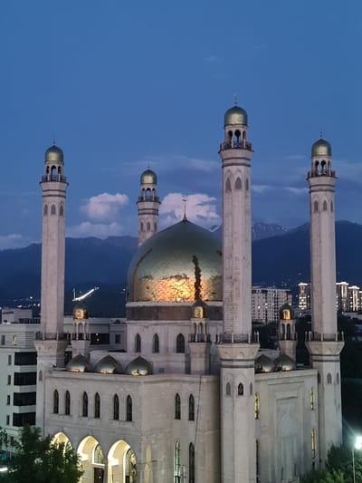 The image shows the Central Mosque of Almaty, Kazakhstan, at dusk. The mosque is a prominent landmark, characterized by its large golden dome and four tall minarets. The architecture is predominantly light-colored stone, with arched windows and balconies visible. The sky is a deep blue, transitioning from twilight to night, with some clouds present. In the background, mountains are visible, their peaks dimly lit. The city lights of Almaty can be seen in the distance, indicating an urban setting. The image captures a serene and majestic view of the mosque against the backdrop of the natural landscape and the city.