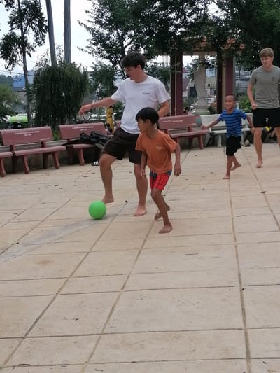 In Da Lat, Vietnam, on a paved courtyard in front of two red concrete benches, four individuals are engaged in a game of soccer. Three are male youths: two appear to be teenagers, one wearing a white t-shirt and dark shorts, and another in a gray t-shirt and dark shorts. The third youth, younger, is wearing a blue and white striped t-shirt and dark shorts. The fourth, a young boy, is wearing an orange t-shirt and red and blue patterned shorts. He is the closest to the viewer, in motion as he kicks a bright green soccer ball. The two teenagers are on either side of him, appearing to participate in the game. The environment includes tall trees, a stone statue, and buildings in the background, suggesting a public park or communal space. The ground is tiled with beige concrete squares.