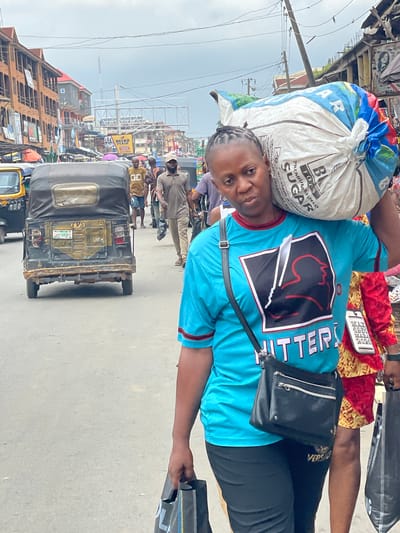 In Fegge, Nigeria, a woman carries a large bag of sugar on her shoulder, walking on a street. She wears a turquoise t-shirt with a red and black graphic and "HITTERS" written on it, along with black pants and a black crossbody bag. Behind her, a yellow auto-rickshaw is parked on the street. The street is lined with buildings and a cloudy sky is visible. Several other people are walking in the background.