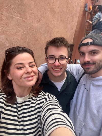 A selfie-style portrait captures three individuals smiling in the foreground against a textured reddish-brown building wall in Marrakesh, Morocco. A female with dark hair and sunglasses on her head, wearing a black and white striped sweater, holds the camera on the left. A male with round glasses, wearing a dark green collared shirt over a white undershirt, is in the center, his right arm around the shoulder of another male on the right. This male wears a grey hoodie and a dark baseball cap backward, displaying a slight beard and stubble. In the upper right background, a male in a blue jacket and dark pants is positioned on a wooden ladder, actively applying material to the wall with a blue-headed tool. The scene depicts a casual group photo with a visible work activity occurring in the background.