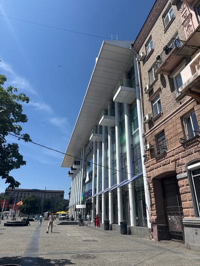 The image depicts a sunny day in a city square in Dnipro, Ukraine. A modern glass-fronted building with white pillars stands on the right, featuring shops on the ground floor. To its right, an older brick building with decorative elements and balconies shows signs of wear. The square is paved with cobblestones and is populated by a few individuals walking, standing, or sitting. A street sign in Cyrillic script indicating "МИРОНОВА" (Mironova) is visible on the older building. In the background, a larger, older building with several stories and flags can be seen across the square. Trees and a blue sky with a few birds in flight complete the scene.