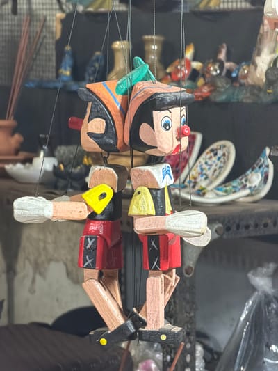 This is a close-up, vertically oriented image of a traditional Indonesian marionette, likely a Wayang Golek puppet. The puppet is made of carved and painted wood.
The subject is a single puppet, seemingly depicted in a shop or market stall. The puppet has a stylized human face with large blue eyes, black eyebrows, and a red nose. It wears a black wig and a headdress or hair ornamentation that is green on one side and orange with blue trim on the other. The puppet's body is segmented, made of cylindrical wooden pieces. Its torso is painted red with black and white decorative elements resembling crossed bands or ribbons. The arms are jointed, made of wood, and extend outwards, with one hand visible with a white glove or wrap. Thin black strings are visible, suggesting the puppet is suspended or ready to be manipulated.
In the background, other objects are blurred and out of focus, including what appear to be more puppets or craft items, as well as shelves and possibly plastic sheeting. The lighting appears to be artificial, typical of an indoor setting. There is no visible text in the image. The overall impression is of a handcrafted item displayed for sale. The location context of Kuta, Indonesia, suggests this puppet is part of the local artisanal crafts and cultural heritage.