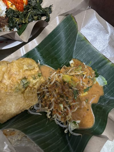 This image shows a close-up overhead view of a meal served on banana leaves and brown paper. The food appears to be a traditional Indonesian dish, possibly Gado-Gado or Pecel, characterized by a mound of mixed vegetables, bean sprouts, and what looks like tempeh or fried fritters, all coated in a rich peanut sauce. The vibrant green of the banana leaf contrasts with the earthy tones of the paper wrapping and the varied colors of the food.
In the foreground, a golden-brown, fried fritter or piece of tempeh is visible, appearing slightly crispy. Beside it, a portion of the vegetable and peanut sauce mixture is prominent, showcasing the textures of the ingredients. In the background, to the left, there's a glimpse of another dish with white rice, dark meat, and bright red chili sauce, also wrapped in paper.
The setting is informal, likely a takeaway meal or a casual dining experience. The lighting suggests it could be daytime, though indoor lighting cannot be ruled out. There are no people visible in the frame. The focus is entirely on the food presentation. The overall impression is one of a hearty and flavorful meal. The location context provided places this meal in Tangerang, Indonesia, which aligns with the style of food presentation and ingredients. No visible text is present in the image.