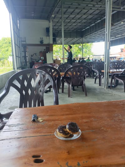 The image depicts an outdoor seating area of a food establishment, likely a roadside cafe or warung, in Lhokseumawe, Indonesia. In the foreground, a weathered wooden table occupies the lower portion of the frame, with two chocolate-frosted donuts on a small glass plate positioned on the right side. A car key with a gold tag is visible on the left side of the table. Behind the table, rows of dark brown plastic chairs are arranged under a corrugated metal roof supported by metal poles. A concrete floor is visible between the chairs and tables. In the background, a person in a black t-shirt is standing, holding a rope or string, and appears to be engaged in some activity or task. Other patrons are seated at tables in the middle ground, some conversing. The background shows a semi-open structure with visible trees and foliage outside, suggesting a rural or semi-urban setting. The overall atmosphere is casual and functional, typical of informal dining spaces in Indonesia.