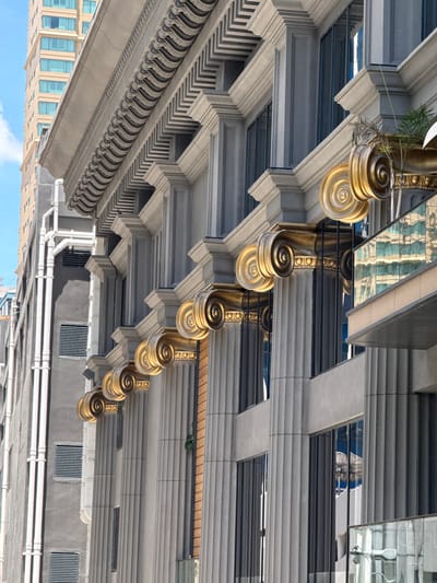 The image shows the facade of a modern building with classical architectural elements, located in Bangkok, Thailand. The building features a series of fluted columns topped with ornate gold volutes, resembling Corinthian capitals. These columns are arranged in a rhythmic pattern along the building's exterior, interspersed with dark windows that reflect the surrounding environment. Above the columns, a decorative cornice with a wave-like pattern runs horizontally. To the left of this facade, another building is visible, taller and constructed with a lighter-colored material, featuring multiple rows of windows. The sky is blue with some scattered clouds. The scene depicts a blend of traditional architectural styles and contemporary building design in an urban setting.