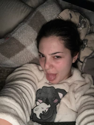 A young woman reclines in bed, her head resting on a plaid pillow. She wears a light-colored, fuzzy pajama top featuring a cartoon cat design. Her dark hair is pulled back, and she looks at the camera with a playful expression, sticking her tongue out. The scene appears to be indoors, softly lit and comfortable. The background includes a patterned blanket or bedding.