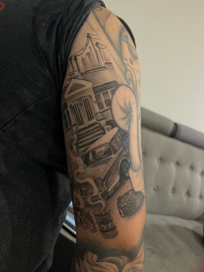 The image is a close-up of a person's arm with a black and grey tattoo. The tattoo depicts a street scene with a building that appears to be a post office or similar establishment with columns and a grand staircase. In front of the building, there is a classic car, possibly a lowrider, with smoke emanating from its exhaust pipes. Below the car, there is a hazardous waste barrel with a biohazard symbol. Further down the arm, a hand is visible, clenched into a fist, with a bag of money on top. The background of the tattoo appears to be abstract or filled with shading. The person is wearing a dark grey t-shirt. The scene is rendered in a realistic tattoo style. The image does not contain any information that would indicate a specific location in Madrid, Spain.