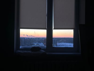 From a dark room, a window reveals a wintry urban landscape at dawn or dusk. Blinds are partially lowered. Buildings and snow-covered ground dominate the view, illuminated by streetlights. A smokestack emits smoke into the colorful sky. On the windowsill, a cushion and a dark object sit. The contrast between the dim interior and the bright outside is striking.
