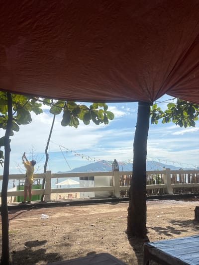 This image depicts a coastal scene, possibly in Đà Nẵng, Vietnam, viewed from beneath a large, reddish-brown tarp. In the foreground, the ground is covered in sandy dirt with scattered debris, including a white plastic bag and what appears to be a disposable cup. Two dark tree trunks are visible, one closer to the left edge of the frame and another more centrally located, with leafy branches extending overhead.
Beyond the trees, a white wooden fence separates a sandy area from the beach and the sea. On the beach, there are a few red chairs and a white umbrella. A person wearing a yellow top and light-colored shorts stands near the fence, holding a mobile phone to take a picture or video. String lights with colorful triangular flags are strung across the scene behind the fence, leading towards a distant island or coastline. The sky is a vibrant blue with scattered white clouds. The overall impression is a casual outdoor setting by the sea, possibly a cafe or rest area.