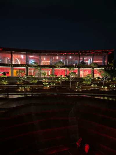 The image depicts a modern, multi-level building with a curved facade illuminated by red lights at night. The building features large glass windows, revealing a red-lit interior with visible architectural elements. In front of the building, there are tiered levels with meticulously sculpted trees and landscape lighting. Several individuals are visible on these tiered levels, appearing to be walking or standing. In the foreground, a dark, possibly water feature, with red lighting is present. In the far right foreground, two individuals are visible, one appearing to be wearing a red garment. The overall scene suggests a public space designed for evening entertainment or gathering. This image is of the "Tsaritsynskiye Baths" complex in Krasnodar, Russia.