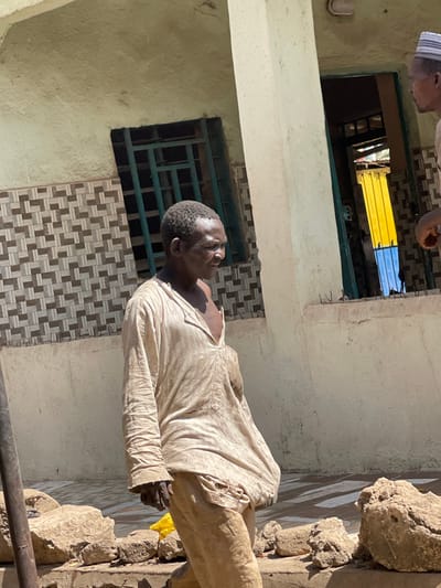 The image depicts an outdoor scene in Zaria, Nigeria, featuring two adult males against a building facade. The primary subject, a male with dark skin and short, dark hair, is positioned slightly right of center, facing right in profile. He wears a light-colored, long-sleeved tunic, open at the neck, revealing his chest, and light-colored trousers. His expression is neutral. To the far right, partially visible, is a second male wearing a light-colored cap and holding a reddish-brown object in his hand.
The background consists of a light-colored building wall with a lower section featuring patterned brown and off-white geometric tiles. Two window or door openings are present: one on the left with dark green metal grilles, and another on the right revealing a vibrant yellow structure and a blue surface deeper inside. A light-colored structural pillar separates the two individuals. The foreground is an unpaved, uneven surface with several large, irregularly shaped earth clods or rocks and a partially visible yellow object. The scene is brightly illuminated by direct sunlight, casting distinct shadows.