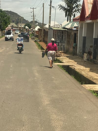The image depicts a street scene in Barakin Akawo, Nigeria. The primary subject is a person walking on the right side of a paved road. The person is wearing a pink shirt and plaid shorts, with a white cap or head covering. They appear to be walking away from the camera, and their shadow is cast on the road.
The setting is a street lined with buildings on the left. Some of these buildings appear to be residential or commercial, with various architectural styles visible, including corrugated metal roofs and brickwork. There is a visible drainage ditch running alongside the road in front of some of the buildings. Power lines and utility poles are also present along the street.
Further down the road, several vehicles are visible, including a black car, a white van carrying goods, and a motorcycle with a rider and a passenger. The motorcycle has some text on its rear panel, but it is not clearly legible. The road itself is asphalt and appears to be in fair condition.
The time of day appears to be daytime, as indicated by the shadows and the lighting. The weather seems to be overcast or hazy, as the sunlight is diffused. The overall atmosphere is that of a typical day in a Nigerian town, with activity on the street and evidence of everyday life. There are no explicit indicators of a specific event, but rather a snapshot of a normal day.