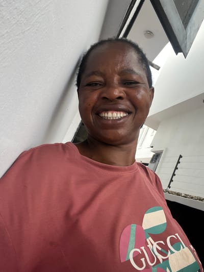 A front-facing, waist-up portrait captures an adult female with dark skin and short, dark hair, smiling broadly with visible teeth, leaning against a white textured wall. She is wearing a salmon-colored short-sleeved t-shirt featuring a multicolored "GUCCI" logo on the chest, designed with geometric shapes and stripes in pink, teal, white, and light brown hues. The background to her right reveals an outdoor setting under bright daylight. Visible elements include a dark, angled overhead architectural beam or roofline, a distant white building, and a dark perimeter fence equipped with security wires or spikes. The scene is located in Lekki, Nigeria.