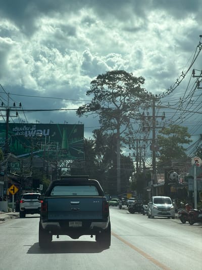 The image captures a street scene in Ko Samui, Thailand, on a cloudy day. The perspective is from inside a vehicle, looking forward. In the foreground, the rear of a dark blue Ford Ranger pickup truck is visible. Ahead of the truck, a white Suzuki Celerio car is driving in the same lane. On the right side of the road, several motorcycles are parked. Further down the street, there are shops and buildings lining both sides of the road, with numerous power lines and utility poles overhead. A large green billboard for Chang beer is prominently displayed on the left side of the road. In the background, a tall tree with a dense canopy stands out against the cloudy sky. The overall impression is that of a typical urban street in a tropical location.