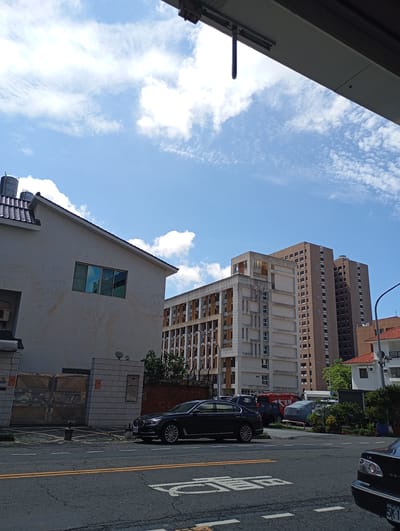 A street-level, full-shot, eye-level perspective shows a clear blue sky with scattered white clouds above a street scene in Kaohsiung, Taiwan. On the left, a light-colored, two-story house with a dark tiled roof occupies the foreground. A metallic gate with intricate patterns is visible in front of the house. To the right of the house, a modern building with a light beige facade and numerous windows extends into the background. Further to the right, two taller, brown apartment buildings stand side-by-side. A portion of a red-tiled roof of a smaller building is visible on the far right. A dark sedan is parked on the right side of the street, and another vehicle is partially visible in the bottom right corner. The street is paved with asphalt and has white and yellow lane markings. In the distance, more buildings and lush green trees suggest an urban environment.