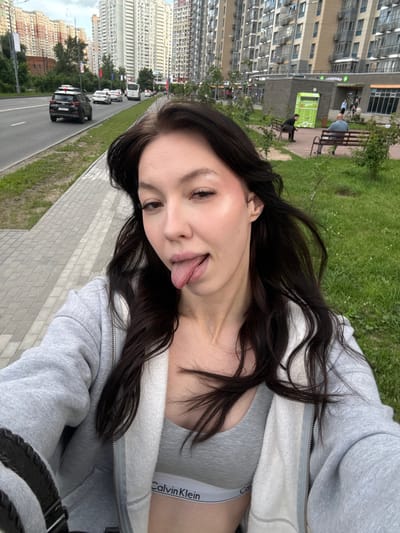 A young woman with long, dark hair is taking a selfie outdoors, sticking her tongue out playfully. She is wearing a grey zip-up hoodie over a grey sports bra with a Calvin Klein logo visible. She is standing on a paved sidewalk next to a grassy area with some trees. In the background, there is a street with cars, and tall apartment buildings typical of the city of Khimki, Russia. The sky is overcast.