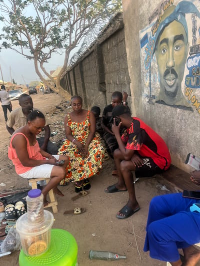 People are sitting and interacting near a painted wall featuring an image of Tupac Shakur and some text in Rido, Nigeria. One group is seated close together near the wall, while another group is seated on stools near an assortment of items, including shoes, containers, and a Pepsi bottle. A tree is overhead, and a barbed wire fence lines a building.