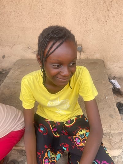 A young person, likely a girl, is captured in a high-angle, outdoor shot, sitting with a bright smile. She has dark skin and her hair is styled in small, neat braids or cornrows, pulled back from her face. She is looking directly at the camera, her expression joyful and engaging.

She is wearing a vivid yellow short-sleeved t-shirt, which appears to have a subtle, light-colored graphic design on the front. Her lower body is clad in dark-colored pants adorned with a lively, colorful pattern of intricate floral or abstract motifs in shades of orange, blue, pink, green, and black. Her arms are resting casually on her legs.

The setting appears to be a casual, outdoor environment, possibly a residential area. The background features a textured, light beige wall on the left side, and rough, grey concrete steps or ground on the right. Some debris, including what looks like a black plastic bag and other scraps, is visible on the steps in the upper right portion of the image. A small, circular, grey object is embedded in the concrete surface behind her head. The lighting is bright and natural, indicating it is daytime. A glimpse of another person's clothing, specifically a white and red garment, is visible at the very bottom edge of the frame, suggesting the presence of someone else nearby. The image is rotated 90 degrees counter-clockwise.