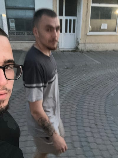 This image captures two males in an urban setting in Lom, Bulgaria, at what appears to be dusk or dawn.
In the foreground, partially visible on the left side of the frame, is a male subject. Only the left side of his face, including his left eye, eyebrow, part of his nose, and a portion of his mouth and chin, is visible. He wears black-rimmed glasses and has dark, short hair and a dark beard or heavy stubble. He is dressed in a dark, possibly black, top or hoodie and appears to be looking upwards and slightly to his right, consistent with taking a selfie or a close-up photograph.
Behind and to the right of the foreground subject, slightly out of focus, stands a second male. He has short, dark hair with closely shaved sides and a longer top. He sports a light mustache and goatee. A small, shiny earring is visible on his left ear. He is wearing a two-toned short-sleeved t-shirt: the upper half is dark grey/charcoal, separated by a thin light grey stripe from the light grey lower half. A dark, possibly black, tattoo is visible on his left forearm. He is also wearing light-colored shorts. His body is angled slightly away from the camera, but his head is turned somewhat towards the viewer.
The ground beneath them is paved with grey interlocking pavers or cobblestones laid in a pattern, with some sparse green vegetation growing in crevices. In the background, there is a light-colored building, possibly beige or light yellow. It features a prominent white door with vertical glass panels on the upper half, and two white-framed windows. The window to the right of the door appears to be slightly ajar, and a white rectangular object or paper is visible inside. A faint artificial light source is visible within the building, illuminating the area around the right window. The overall lighting is low, contributing to a muted color palette.