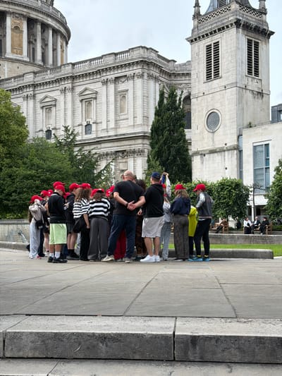 The image depicts an outdoor scene in the city of London, United Kingdom, featuring St. Paul's Cathedral and surrounding architecture. In the foreground and midground, a group of approximately 15-20 individuals is gathered on a paved stone area. Many of the younger individuals in the group are wearing red baseball caps and casual attire such as t-shirts, shorts, and hoodies. An adult male in a black t-shirt and dark pants stands with his back to the viewer, appearing to address or interact with the group. Another adult male in a black t-shirt and white shorts stands to the right, holding a blue bottle and a phone, seemingly taking a picture or video. Several other adults are interspersed within the group.
The background is dominated by architectural structures. St. Paul's Cathedral's classical stone facade, featuring columns and a domed structure, occupies the upper left portion of the image. To its right, a prominent stone clock tower or steeple with louvered vents and a round window is visible. Further to the right, a modern stone building with large windows is present. Between these structures and the group of people, there are several large green trees and a manicured grassy area with stone benches where a few people are seated. The sky is overcast. The ground in the immediate foreground consists of rough-hewn stone steps leading up to the large, grey flagstone pavement where the group stands. A water feature is partially visible on the far left.