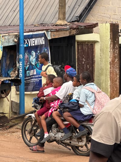 In Ihiagwa, Nigeria, several children are crammed onto a motorcycle parked along a weathered street. The children wear various casual and school-style clothes. A blue utility pipe, marked "13 NDDC", runs horizontally across the lower left of the frame, in front of a weathered wall and a sign advertising "X Viewing" for live matches and shows. The street appears gritty and somewhat unkempt, with rusted metal elements visible.