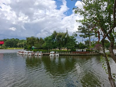 This image depicts Xuan Huong Lake in Da Lat, Vietnam, on a cloudy day. The foreground is dominated by the dark, rippling water of the lake, reflecting the overcast sky. Along the shoreline, a row of small, white paddle boats with orange life preservers are moored. Behind the boats, a white picket fence separates the water from a lush green area with trees and bushes. A sign with white lettering on a dark background reads "THUY TA RESTAURANT & COFFEE," indicating a commercial establishment. In the distance, more trees and vegetation are visible, and the sky is filled with dramatic, textured clouds. To the far left, a red and white sign with "AUS" is partially visible, possibly advertising a business or event. No people are clearly discernible in the scene.