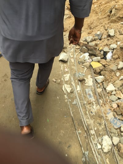 A person in gray attire is seen from the torso down, walking on a concrete surface with debris. The area is scattered with rocks and bits of trash near what appears to be metal rods. The person is wearing brown sandals, and a silver bracelet is visible on their left wrist. The view is obstructed by a blurry object in the foreground.