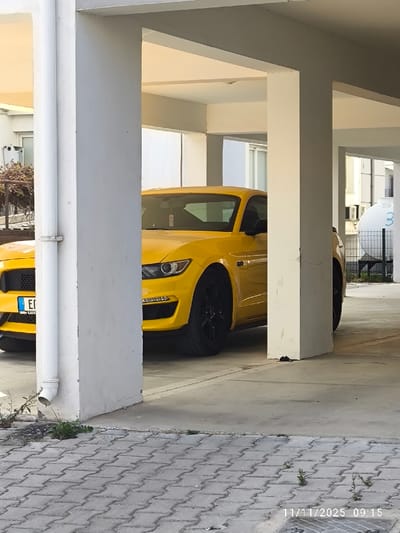 A yellow Ford Mustang is partially visible under a concrete carport structure. The car's front end and side are seen between the support pillars. The car has black wheels. The ground is paved with gray bricks. There is a timestamp on the bottom right corner of the image.