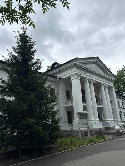 The image depicts the Vnukovo Children's Railway building in Vnukovo, Russia, on a cloudy day. The building is a white, neoclassical structure with a prominent portico supported by large, fluted columns. The facade features intricate decorative elements, including a pediment with a circular emblem and detailed moldings. Several balconies with black railings are visible on the upper level. To the left of the building, a tall, dark green spruce tree dominates the foreground, its branches extending upwards towards the overcast sky. The ground in front of the building consists of a paved pathway and a grassy area with some weeds and small plants. A signpost with information and a large white sphere are situated near the entrance. The overall scene is serene and architectural, with a focus on the grandeur of the building and the natural element of the tree.
