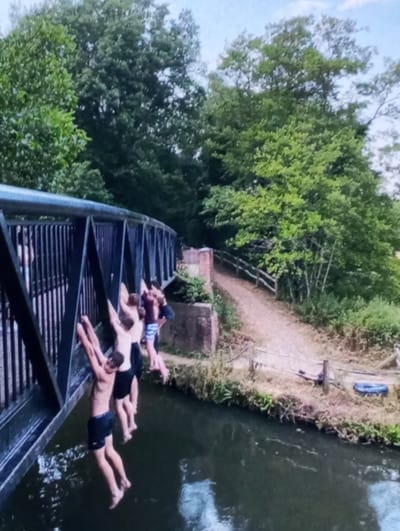 The image displays a daytime scene where four young males are in the process of jumping from a dark-colored metal pedestrian bridge into a body of water, identified as a canal or river in Brzezina, Poland. The bridge features a visible lattice or truss structure on its sides, from which the individuals are suspended.

The closest person on the left, a young male, hangs with arms extended upwards, gripping a vertical bar of the bridge's side structure, wearing dark swim shorts. His feet are clear of the water, suggesting an active jump or preparation to drop. To his right, three other young males are similarly suspended from various points of the bridge framework. One wears dark shorts, and the individual furthest right wears patterned shorts. Their bodies are positioned above the water, indicating recreational activity.

The water beneath the bridge is dark and appears calm, reflecting portions of the sky and surrounding trees. On the far bank, a dirt or gravel path runs alongside the water, marked by a multi-rail wooden fence. Dense green deciduous trees form a lush background, suggesting a heavily vegetated area. A brick structure, possibly a bridge abutment, is visible on the far bank directly beneath the bridge. A blue inflatable ring is discarded on the ground near the water's edge on the far bank. The sky is bright, indicative of daylight conditions.