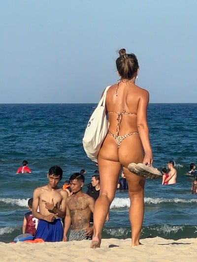 An overhead shot shows a woman from behind, wearing a leopard-print bikini and holding a pair of shoes in her right hand, as she stands on a sandy beach with the ocean in the background. A white tote bag hangs from her left shoulder. In the shallow water, several people are visible, including two men in the foreground standing waist-deep in the ocean, one wearing blue shorts and the other with patterned shorts. A person in a red top is further out in the water. The scene is set on a beach in Đà Nẵng, Vietnam, indicated by the presence of local individuals and the general ambiance of a popular tourist destination. The sky is clear and blue, and the ocean has moderate waves.