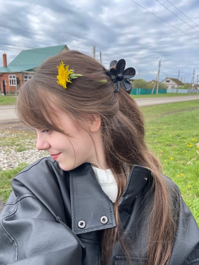 The image shows a young woman in an outdoor setting. She has long brown hair, adorned with a yellow dandelion near her forehead and a black flower clip towards the back of her head. She is wearing a black leather jacket over a white top. The background indicates a rural area, with a gravel road, grass, and buildings visible. The sky is overcast. The location is near Karmaskaly, Russia, based on the presence of a residential building and utility poles in the area.