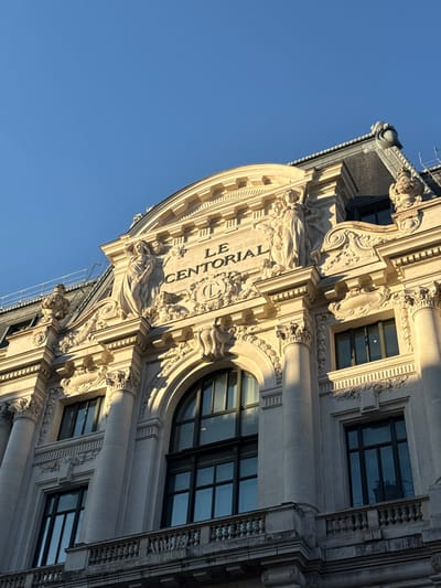 The image displays an upward view of the ornate limestone facade of the Le Centorial building, situated at 33 Rue de Miromesnil, 75008 Paris, France. The building exhibits a Neoclassical architectural style with extensive decorative elements. Centrally located on the upper visible floor is the inscription "LE CENTORIAL" in capital letters. This inscription is flanked by two large, draped allegorical female sculptures standing on pedestals, with additional smaller bas-reliefs below them. The facade features fluted columns with ornate Corinthian capitals, supporting an elaborate entablature and cornice work. Multiple large windows are visible, including a prominent arched window and several rectangular multi-paned windows. Reflections are present in the window panes, and interior venetian blinds are partially visible through some. A stone balustrade with multiple balusters runs across a lower section of the building. Portions of a dark mansard roof with decorative cresting are visible at the top of the structure. Strong, directional sunlight illuminates the building from the left, casting deep shadows that emphasize the architectural contours and sculptural details. The sky above is clear and uniformly blue. No individuals, actions, or specific interactions are depicted.