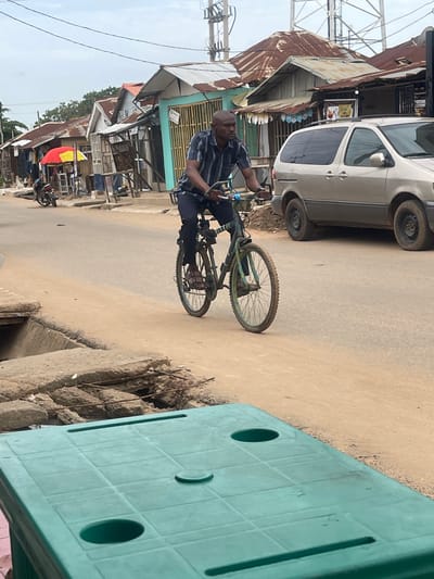 A man rides a bicycle down a dusty street in Abuja, Nigeria. He wears a blue and white striped shirt and dark pants, with sandals on his feet. The bicycle is green and appears to be well-used, with knobby tires. In the background, a row of small shops with corrugated metal roofs lines the street. A silver minivan is parked to the right of the bicycle. The sky is overcast, and the overall atmosphere of the scene is one of everyday life in a busy urban environment. The foreground is dominated by a green plastic table.