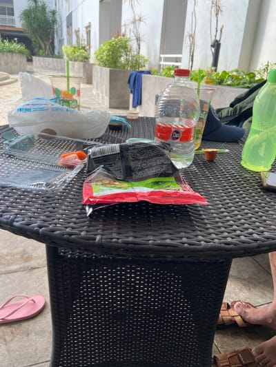 A rattan table is littered with personal belongings. A red and white water bottle, a green bottle, a bag of chips, a cup, a takeout container, and a blue sock are on the table. In the background, there is some greenery, a building, and stairs. A pair of sandals and bare feet are visible on the upper right side of the image. A pink flip-flop is on the floor next to the table.