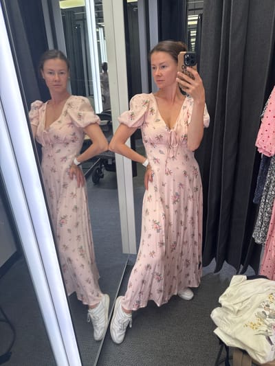 This is a vertical, medium shot of a woman taking a mirror selfie in what appears to be a clothing store fitting room. The woman is wearing a light pink, floral-print midi dress with puff sleeves and a tie-front bodice. She has her hair pulled back and is looking at her phone, which she holds up to take the picture. She is also wearing white sneakers. Her reflection is visible in a large mirror in front of her, showing her full outfit and the fitting room.

The fitting room has a dark grey carpet and dark grey curtains visible in the background. There are also clothes hanging on a rack behind her, including a pink dress with white polka dots and a white t-shirt with a floral design. The lighting is bright and appears to be from overhead fluorescent lights, suggesting it is daytime. The woman has a neutral expression on her face. There is no visible text in the image. The location context provided is Riga, Latvia.