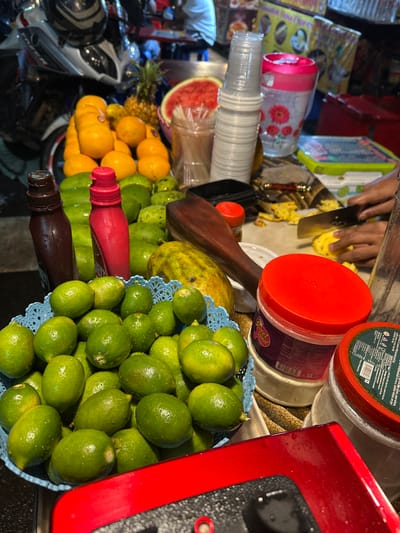 The scene depicts a street food stall in Dhaka, Bangladesh, captured at night under artificial illumination. The primary focus is on a display of fresh fruits and preparation activity.

In the immediate foreground, a blue plastic basket holds a quantity of wet, green limes. To its right, a partial view of a red surface, possibly a counter or machine, is visible. Behind the limes, two upright squeeze bottles are present: one dark brown and one red. A large mound of additional green, round fruits is situated behind these bottles.

Central to the image is a vibrant array of fruits: a substantial pile of bright orange citrus fruits (likely oranges or mandarins), a whole pineapple, and a large slice of watermelon with its red flesh exposed.

To the right, a stack of clear plastic cups is visible, alongside a pink and white floral-patterned drinking bottle. A wooden pestle or similar tool rests on the surface. Multiple plastic containers with red lids are present, with one container clearly labeled "Sweet Land."

In the upper right, a pair of human hands is actively engaged in slicing a yellow fruit, identified as a pineapple, on a cutting surface with a silver knife. Smaller, already cut pieces of pineapple are scattered nearby.

The background on the left reveals the front section of a parked motorcycle with a dark helmet placed on its seat. The overall environment suggests an outdoor market or street vendor setup, characterized by various objects and equipment common to such operations, illuminated by overhead lights. The ground or immediate surface appears dark.