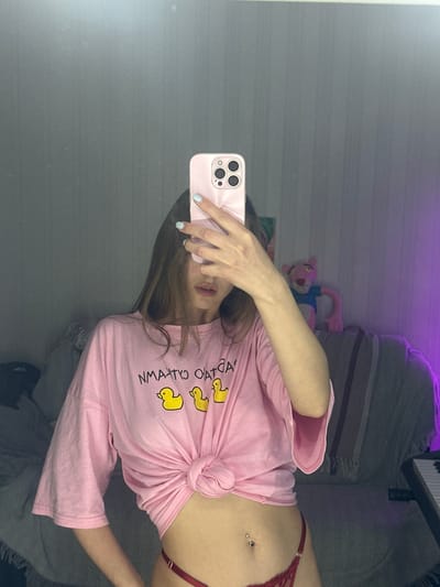 A young woman, with long brown hair, is reflected in a mirror. She is wearing a pink t-shirt with a knot tied at the waist, which exposes her midriff. The t-shirt has a graphic of three yellow ducks. She is also wearing red lace underwear. Her left hand is holding a pink phone in front of her face, obscuring it. She has light blue nail polish on her fingernails, and a belly button piercing. In the background, on a couch, a pink panther stuffed animal can be seen. There is also a keyboard on the right side of the frame. The room is dimly lit with a purple light illuminating the right side of the background. The setting appears to be an indoor residential space.