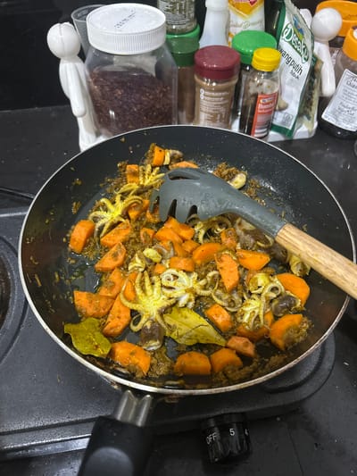The image shows a close-up view of a black frying pan on a black stovetop, filled with a dish containing small octopuses and chopped carrots in a yellowish-brown sauce. A grey and wood-handled pasta server is resting in the pan. In the background, several bottles and jars of spices and condiments are visible on a countertop. One bottle on the far left contains reddish-brown granular material. To the right of that, a clear plastic jar with a white lid is filled with a similar granular substance. Other bottles include ones with green lids, red lids, and yellow lids. A white packet with green writing is also present. The stovetop has a control knob labeled "OFF", "5", and "4". The overall environment appears to be a kitchen. There are no people visible in the image.