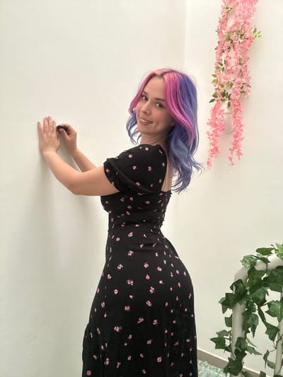 This media file features a young woman posing against a plain white background. The image is oriented such that she appears to be leaning diagonally on a vertical surface, her head positioned towards the top-left and feet towards the bottom-right. She has distinctive hair dyed in shades of purple and pink and is wearing a black dress with a pattern of small pink flowers. Her facial expression is joyful, with a wide, open-mouthed smile directed towards the camera. She rests on her bent forearms and hands, with one hand grasping a small, dark, cylindrical object.
Above her head, stretched horizontally across the white background, is a decorative garland of artificial pink flowers, resembling wisteria or cherry blossoms. To the right of this, another garland of artificial green ivy is similarly draped. The setting appears to be a clean, minimalist indoor space, likely a room with white walls or a ceiling, which provides a stark contrast to the colorful elements of the subject and decorations. There is no visible text within the image. While the location context is provided as the City of Zagreb, Croatia, there are no specific visual cues within the photo to indicate this. The overall impression is one of playfulness and artistic perspective.