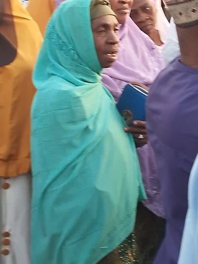 A woman in a light blue headscarf and matching shawl stands among a crowd of people. She has dark skin and is holding a blue book with a silver design. Other individuals are visible, including one in a lavender headscarf and another in an orange top. The person to her right wears a purple shirt and the person to the left has a white garment with brown buttons.