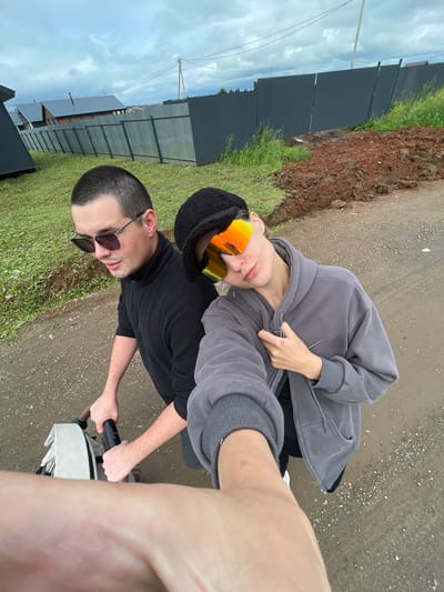Two individuals are captured in a selfie, likely in the village of Podshivalovo, Russia, as indicated by context. The person on the left, wearing sunglasses and a black turtleneck, is positioned behind what appears to be a stroller. To the right, another person, sporting a black cap and bright orange-tinted sunglasses, leans back, holding the selfie arm. They are dressed in a gray zip-up hoodie. The background shows a suburban or rural setting with houses, a dark metal fence, a patch of disturbed earth, and a cloudy sky. The ground is a dirt or gravel path.