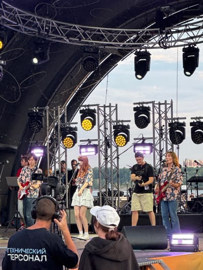 On an outdoor stage in Novosibirsk, Russia, a musical performance is underway. The stage is equipped with multiple lighting rigs and sound equipment, including large speakers. A band, consisting of at least three musicians, is performing. One musician is playing a red electric guitar and singing into a microphone. Another musician, in a floral shirt, is also playing an electric guitar. A third musician, wearing a white skirt and floral top, is singing. Behind the band, a cameraman with a professional video camera is filming the performance. In the foreground, a person wearing headphones and a black t-shirt with the Russian text "ТЕХНИЧЕСКИЙ ПЕРСОНАЛ" (Technical Personnel) is visible, indicating their role in the event. Another person in a white hat is partially visible in the lower right corner. The sky above is overcast, suggesting a cloudy day. The overall scene depicts a live music event, possibly a festival or concert, taking place in an urban outdoor setting.
