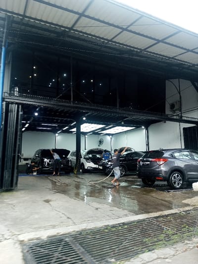 The image captures a bustling car wash facility, likely an indoor or covered multi-bay station, with several vehicles being cleaned by workers. The scene appears to be daytime, though the well-lit interior suggests artificial lighting is primarily illuminating the work area.
The facility has at least two levels. The lower level serves as the car wash area, featuring a concrete floor that is visibly wet from water and cleaning activities. Drainage grates are embedded in the concrete in the foreground and towards the right. Overhead, a corrugated metal roof provides shelter, supported by a metal framework. The bays are brightly lit by numerous fluorescent or LED lights mounted on the ceiling and walls. Large industrial fans are also visible on the back wall, suggesting ventilation.
Three men, presumably car wash employees, are actively engaged in cleaning vehicles. In the center, a man wearing a dark t-shirt and shorts is holding a white hose, spraying water towards the cars. To his left, another man in a dark t-shirt and long pants is bending down near the front driver's side wheel of a dark SUV, appearing to scrub or rinse the lower portion of the vehicle. A third person is partially visible further to the left, also bent over.
Several cars are in various stages of being washed. On the far left, a dark SUV, possibly a Hyundai Palisade, has its hood open and is being worked on. In the middle, a white sedan or SUV also has its hood open, indicating a more thorough cleaning process, potentially including the engine bay. On the far right, a dark gray SUV, which resembles a Honda HR-V, is parked, with its rear facing the viewer. Its license plate is partially visible and appears to read "B 1254 TYY" or a similar combination of letters and numbers. Another dark car is partially visible behind the white vehicle.
The upper level of the building features large, dark-tinted glass windows that reflect interior lights, suggesting it might house offices, a waiting area, or storage. The overall structure is made of metal, with dark beams and supports. On the exterior wall of the upper floor, to the right, an air conditioning unit is mounted. A blue structural pillar is visible on the far left, and a partially open accordion-style metal gate or door is also seen on the left side of the frame. The overall impression is one of an active and well-equipped professional car wash establishment.