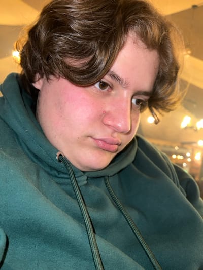 A person with light skin and brown hair wearing a dark green hoodie looks towards the upper left corner. The hair has a loose, wavy texture, and a few strands fall towards the forehead. The face is round with full lips and a slightly upturned nose. The hoodie's drawstring is visible near the neck. Soft lighting illuminates the skin, creating subtle shadows and highlights.