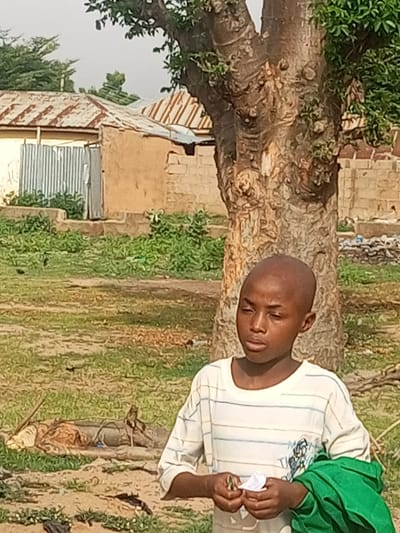 The image depicts a young boy with a shaved head standing in an outdoor environment. He is wearing a striped t-shirt and holding a green cloth in his right arm. In his left hand, he is holding a small white object. Behind him, a large, textured tree trunk dominates the frame. In the background, traditional Nigerian architecture is visible, with buildings featuring corrugated metal roofs and mud or brick walls. The ground is a mix of dirt and sparse vegetation. The scene appears to be in a residential area of Gombe, Nigeria, characterized by its rustic buildings and natural elements.