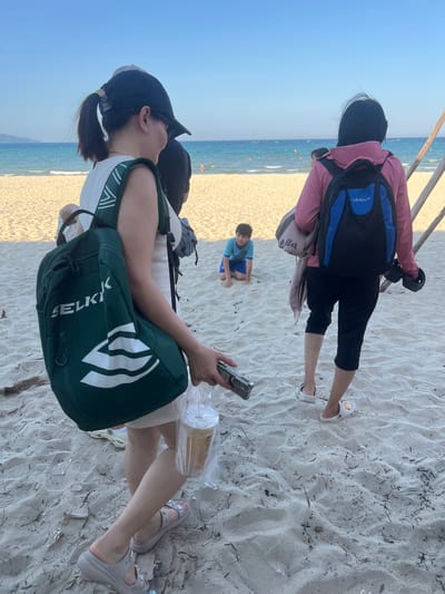 The image shows a group of people on a sandy beach in Da Nang, Vietnam, with the ocean and sky in the background. In the foreground, a woman wearing a black baseball cap and a white top is carrying a green backpack. She is holding a plastic bag with a drink. To her right, another person wearing a pink jacket and a blue and black backpack stands facing away from the camera. Further back on the beach, a young boy in a blue shirt and shorts is kneeling in the sand. The scene is illuminated by natural daylight, suggesting it is daytime.