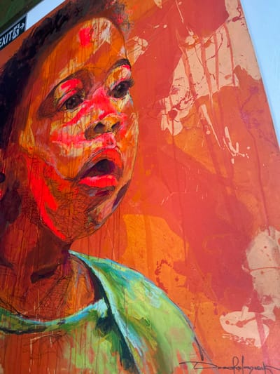 This image depicts a large, vertically oriented, expressive mixed-media portrait painting, prominently featuring the head and upper torso of an individual, likely a child, rendered in a bold, abstract style. The subject's skin tones are predominantly orange and red, with striking neon pink and red highlights applied around the eyes, nose, and open mouth, suggesting an intense or surprised expression with the gaze directed upwards and slightly to the right. Dark hair is visible along the left side of the head. A bright green garment with blue accents is depicted on the subject's left shoulder and chest. The painting's background consists of abstract orange and red fields with visible paint drips, splatters, and textured applications. A partial artist's signature is visible in the bottom right corner of the artwork. The painting is mounted on a plain, light-colored wall. In the upper left corner of the frame, a white rectangular sign with a green border is partially visible, displaying the word "EXIT" in white capital letters accompanied by a black arrow pointing to the right. The scene is located in Alaguntan, Nigeria.